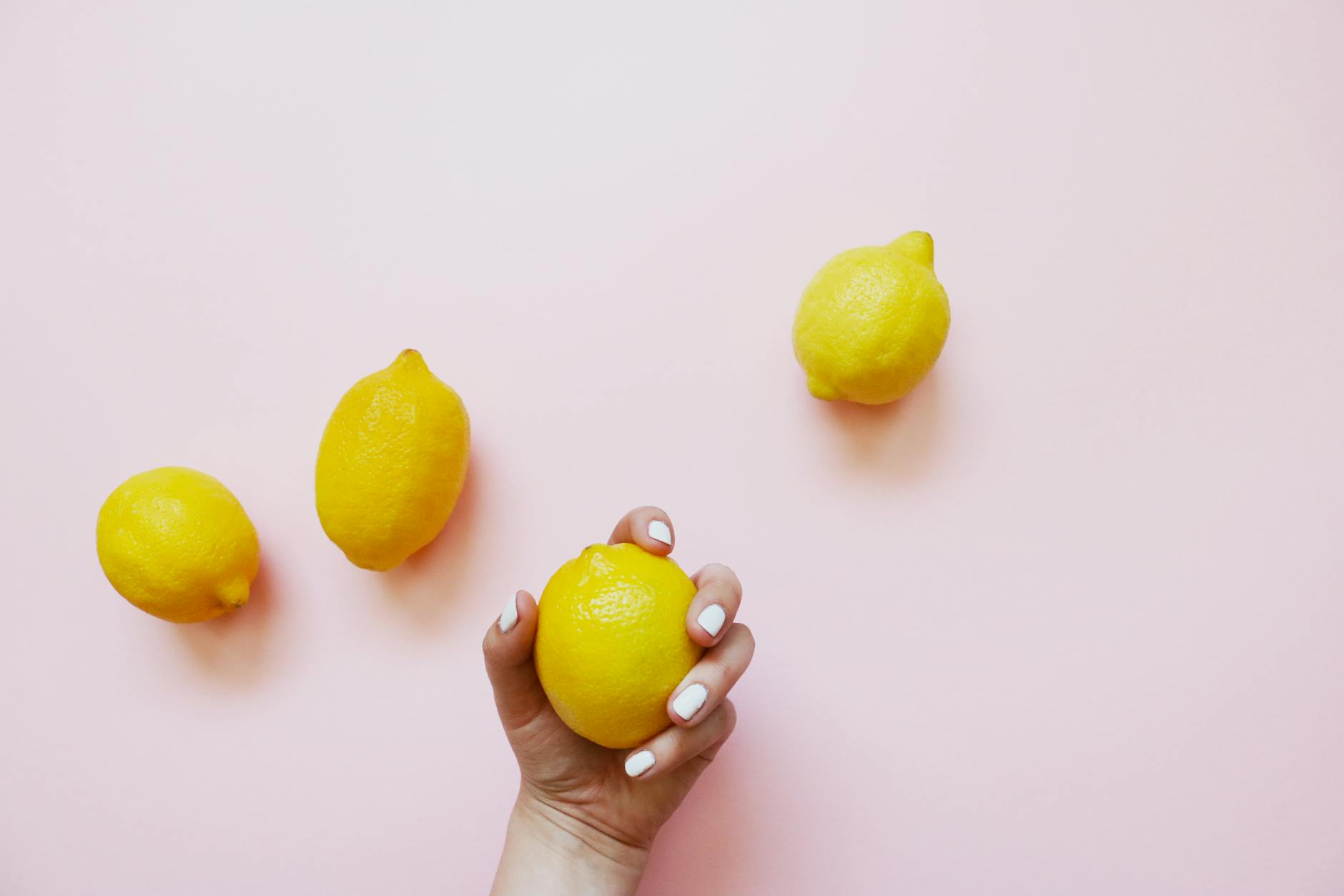 Ruka s bílými nehty drží citron na měkkém růžovém pozadí obklopeném třemi dalšími citrony.