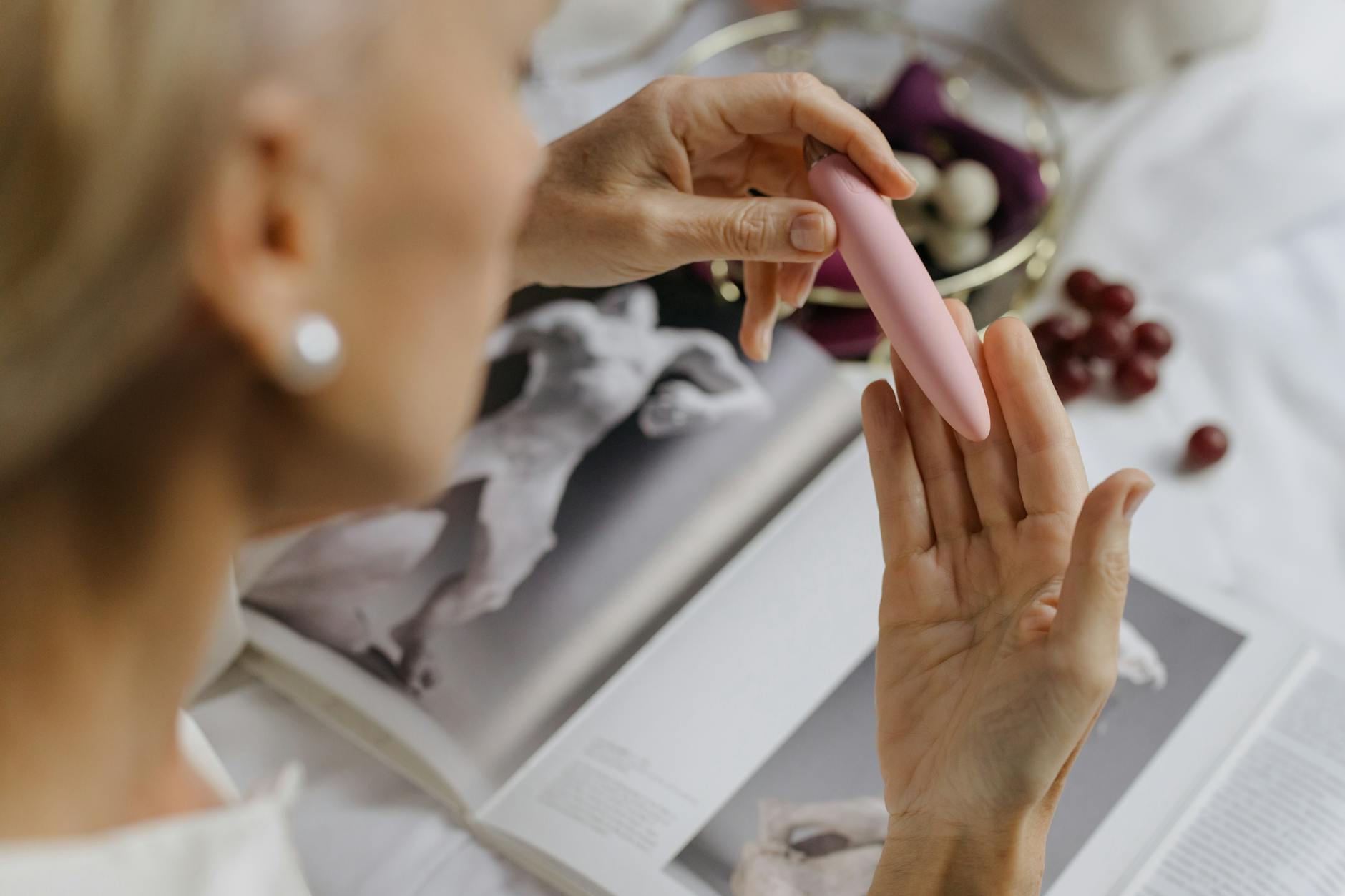 Blízký pohled na ruku držící růžový vibrator se speciálními uměleckými knihami v pozadí, zdůrazňující intimní a osobní téma