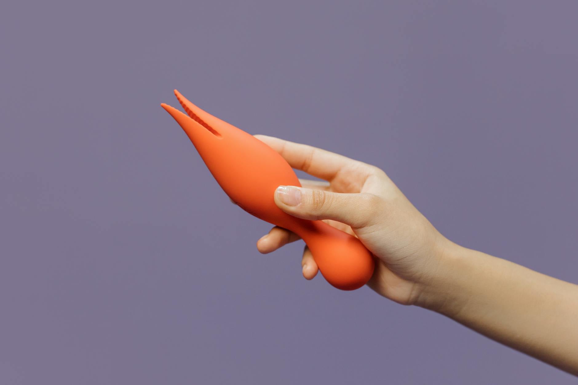Ruka držící moderní oranžový vibrator proti minimalistické fialové pozadí, symbolizující zvýšení intimity v dlouhodobém vztahu